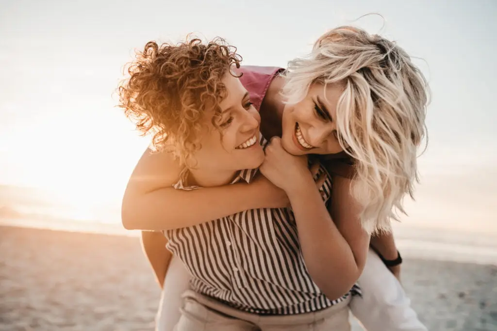Laughing young woman giving her girlfriend a piggyback while having fun together at a beach at sunset
