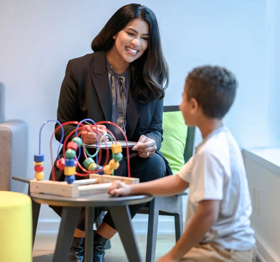 A female therapist sits in a chair beside her young patient as they play together with a bead maze. The therapist is dressed professionally in a suit and has a tablet out on her lap to take notes about the session. The young causally dressed boy is kneeling on the floor n front of the toy as they talk together.