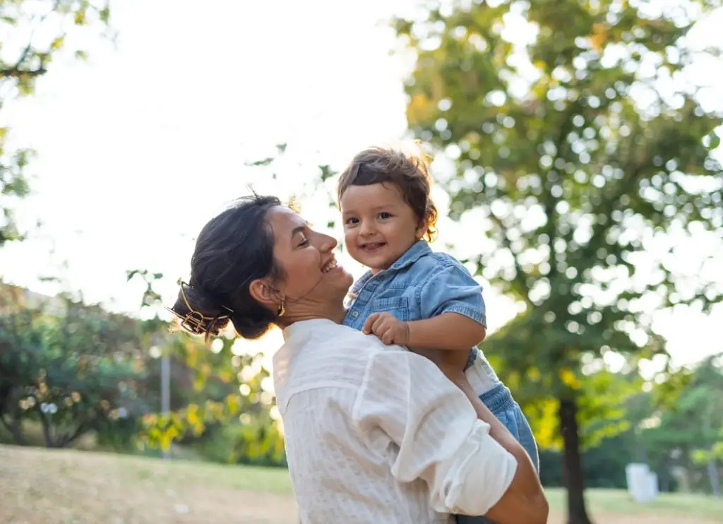 A smiling mother lifts her happy child in a sunny park, surrounded by lush green trees. The moment captures the joy and warmth of a family day outdoors.