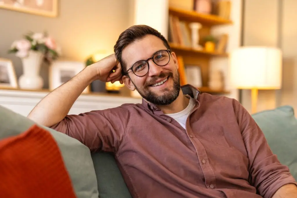 A happy bearded man wearing glasses and a casual shirt smiles in a softly lit home interior. His friendly and confident expression creates a warm, welcoming atmosphere